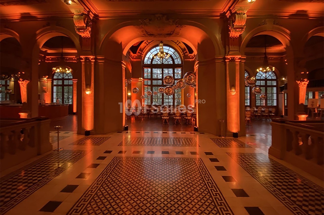 Salle élégante avec arches, sols carrelés et éclairage orange, tables alignées devant de grandes fenêtres.