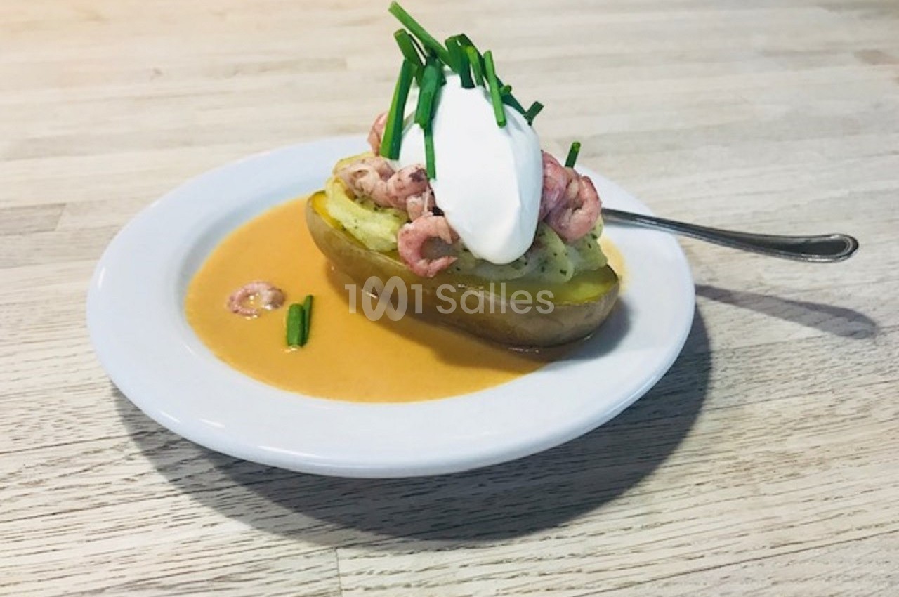 Pomme de terre garnie de crevettes, crème fraîche et ciboulette, servie avec une sauce sur une assiette blanche.