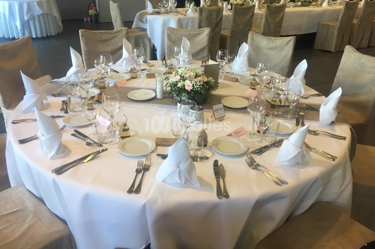 Table ronde élégamment dressée avec nappes blanches, serviettes pliées, vaisselle et centre de table floral.