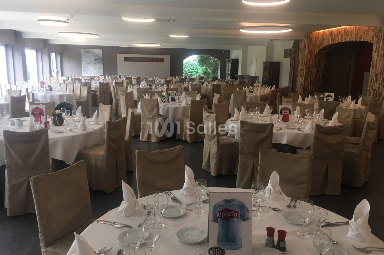Salle de réception avec tables rondes dressées, nappes blanches, serviettes pliées et chaises couvertes de housses beiges.