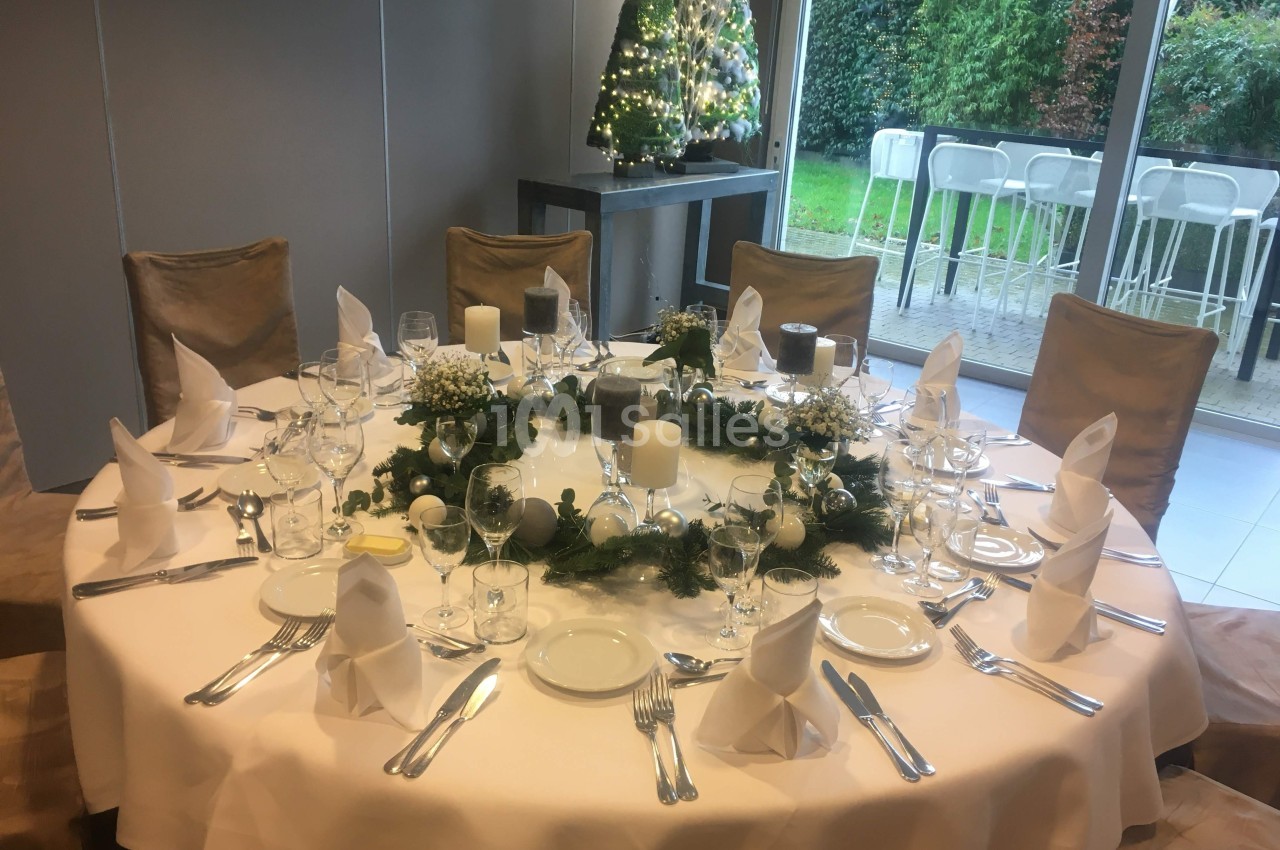 Table ronde élégamment dressée avec vaisselle, serviettes pliées et décoration florale, près d'une baie vitrée et d'un sapin…