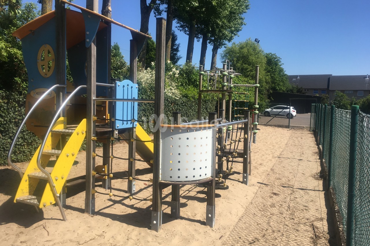 Structure de jeux en plein air avec toboggan et modules d'escalade, installée sur un sol sablonneux près d'une clôture.