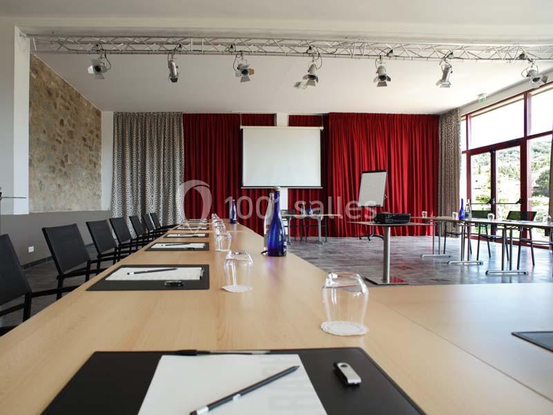 Salle de réunion lumineuse avec tables disposées en U, écran de projection, rideaux rouges et matériel de présentation.