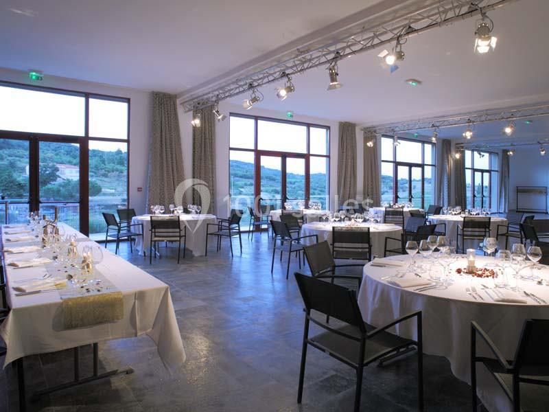 Salle lumineuse avec de grandes baies vitrées, tables rondes et rectangulaires dressées pour un événement.