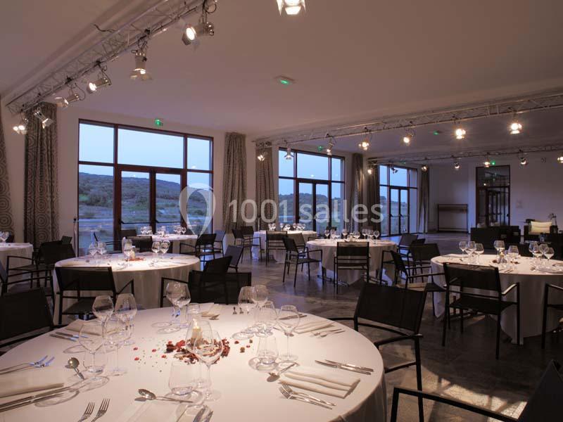 Salle de réception spacieuse avec tables rondes dressées, grandes baies vitrées et vue sur un paysage naturel.