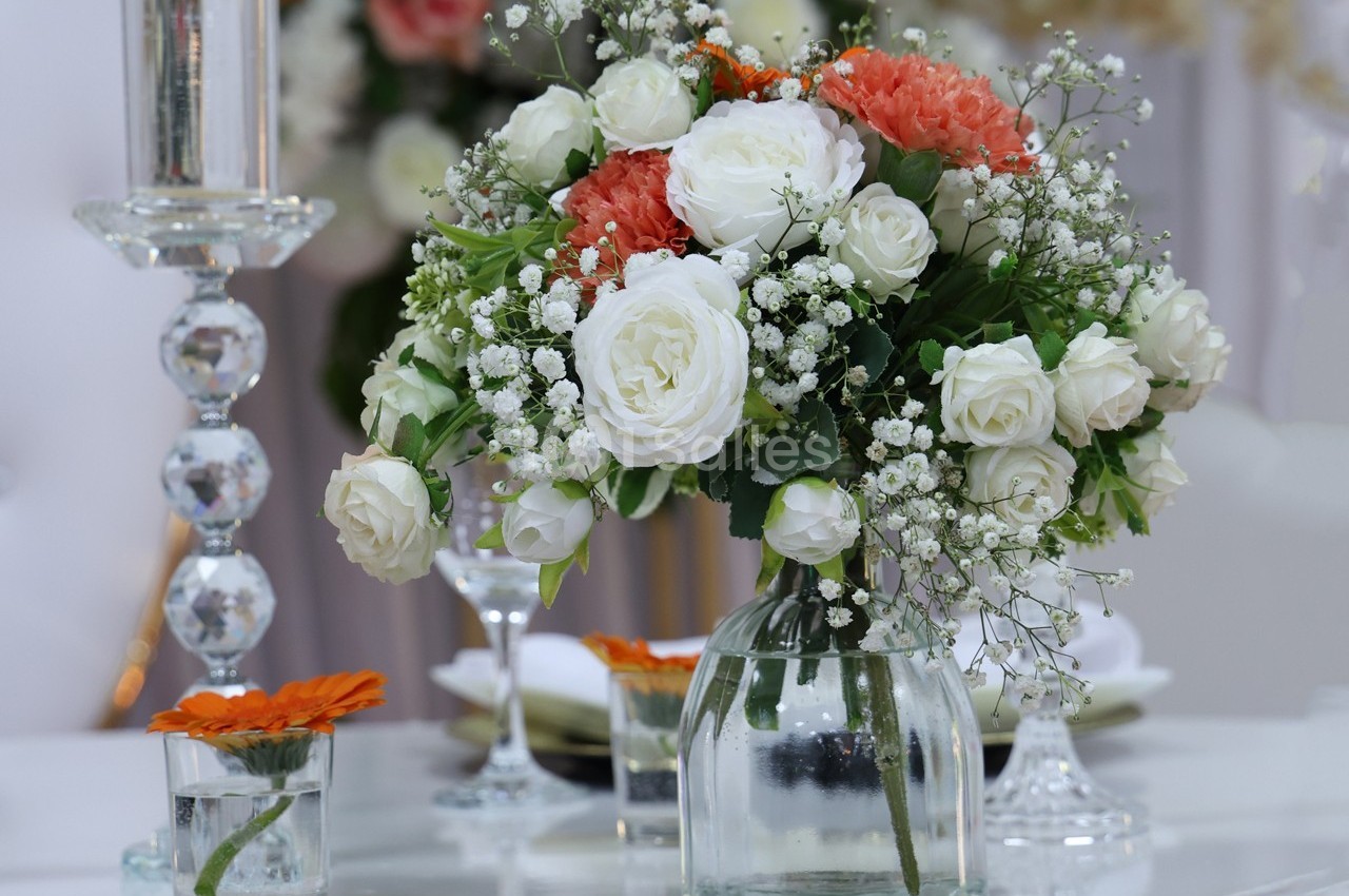 Bouquet de roses blanches et fleurs orange dans un vase en verre, posé sur une table décorée avec des bougies et des verres.
