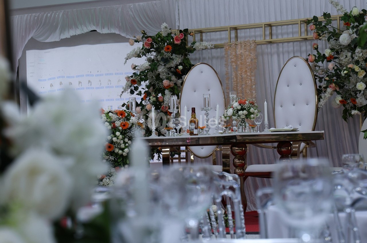 Table décorée pour un mariage avec chaises élégantes, fleurs blanches et orangées, et bougies sur fond de rideaux.