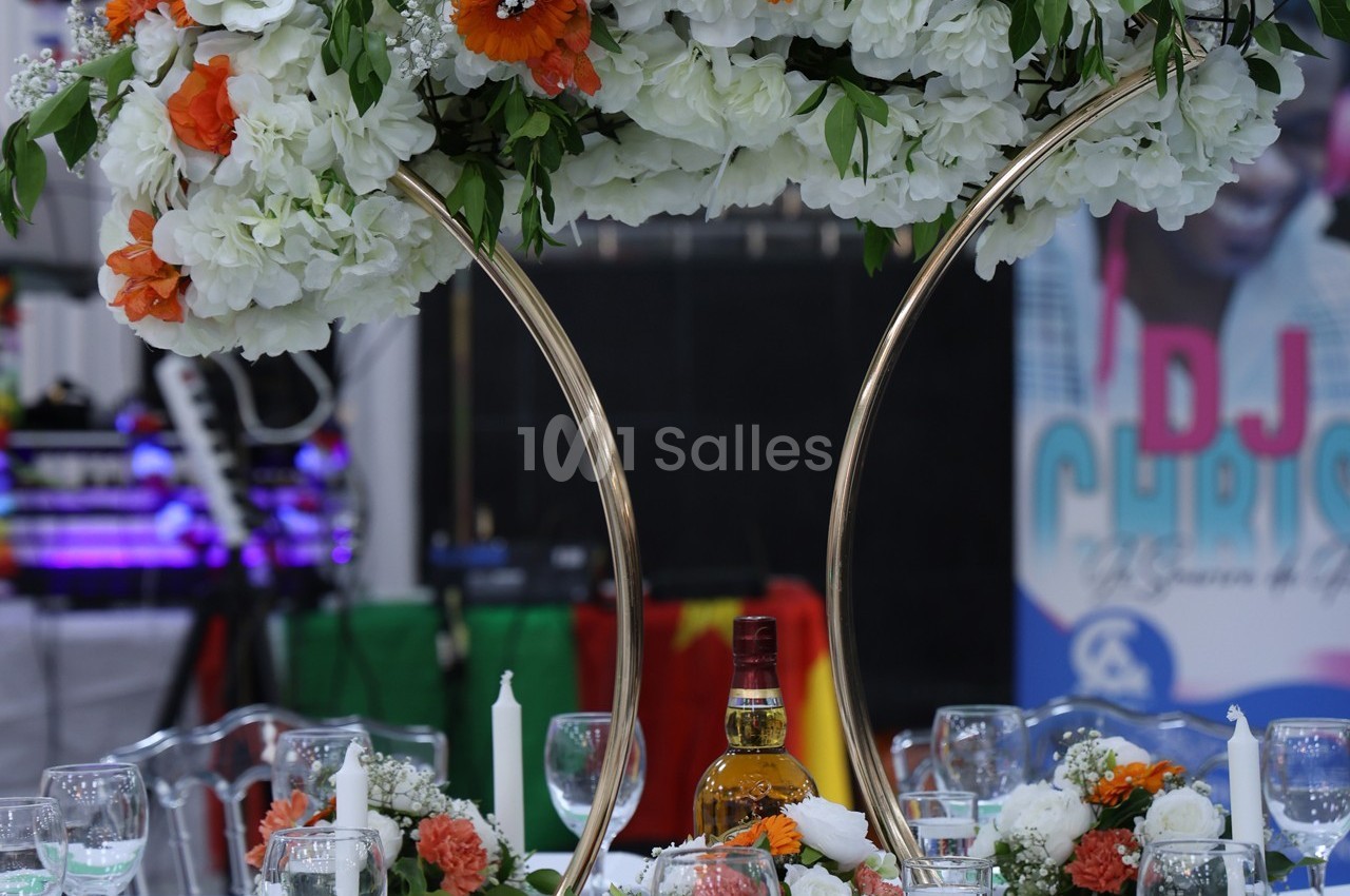 Centre de table floral avec des fleurs blanches et orange, entouré de verres, bougies et une bouteille au centre.