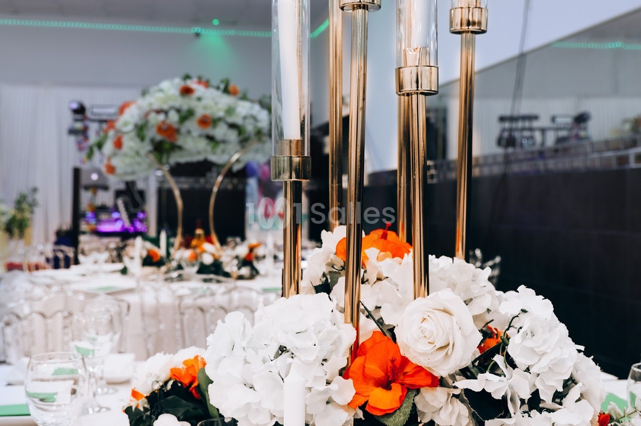 Centre de table floral avec roses blanches et fleurs orange, entouré de chandeliers dorés dans une salle de réception.