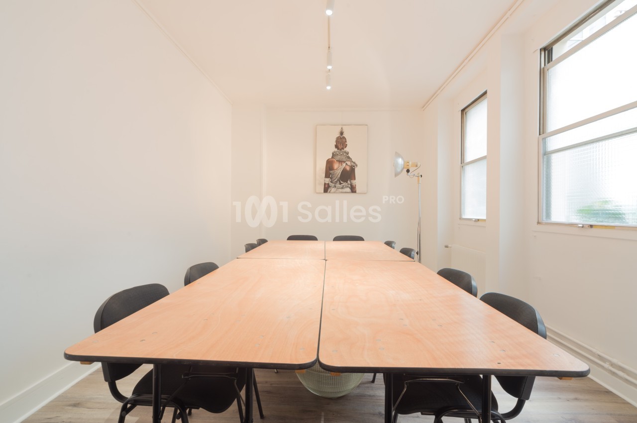 Salle de réunion lumineuse avec une grande table en bois entourée de chaises noires et un tableau mural au fond.
