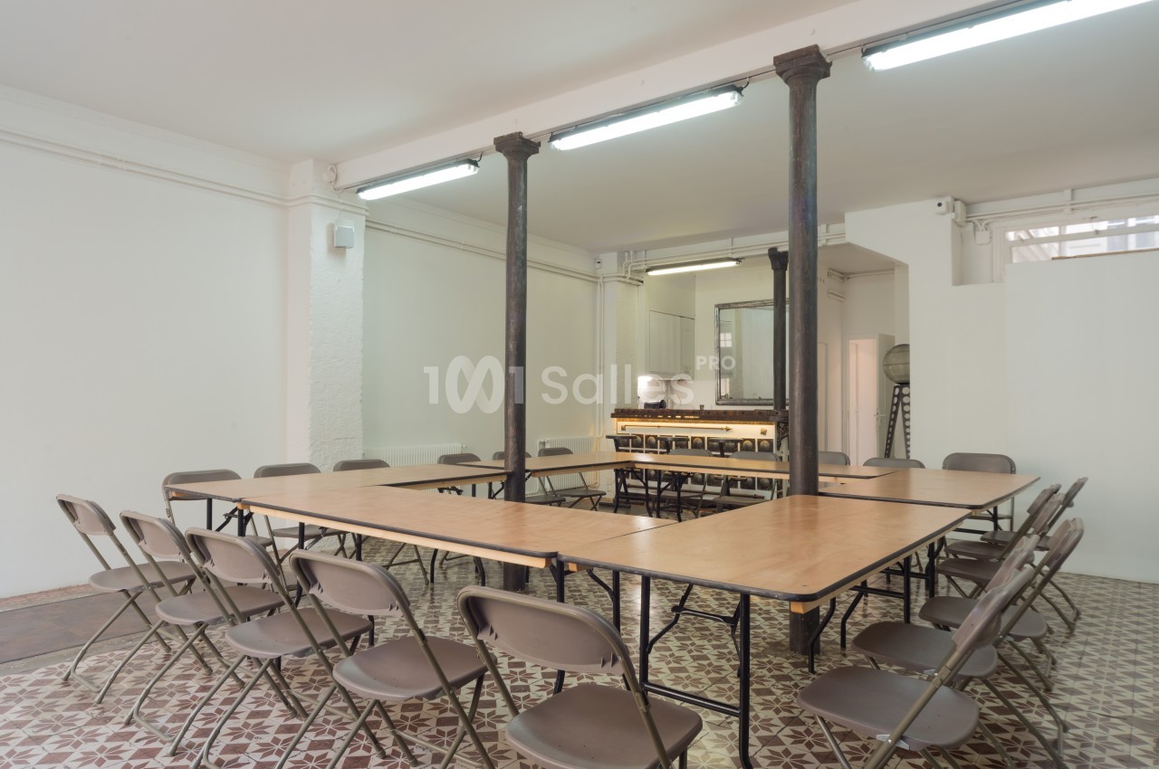 Salle lumineuse avec tables en U, chaises pliantes, colonnes métalliques et sol en carreaux de ciment.