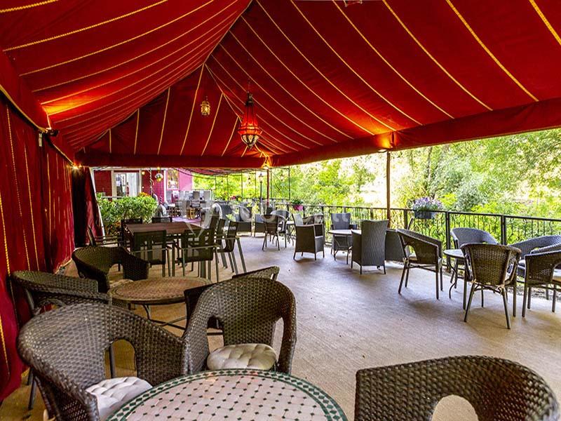 Terrasse couverte avec des tables et chaises en osier, entourée de verdure et éclairée par des lampes suspendues.