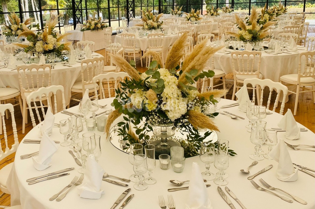 Salle de réception décorée avec des tables rondes, nappes blanches, centres de table floraux et chaises blanches.