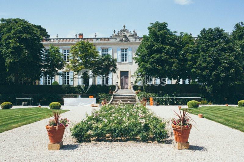 Façade d'un grand bâtiment historique entouré d'arbres, avec un jardin aménagé et des allées gravillonnées au premier plan.