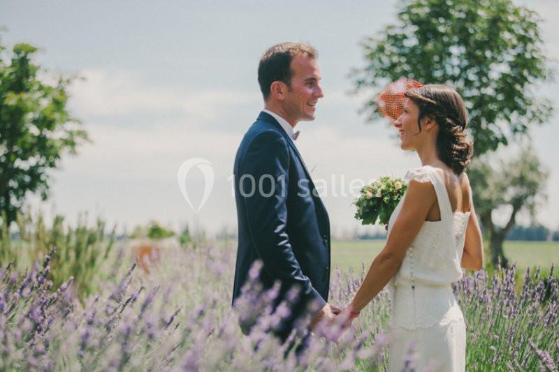 Un couple en tenue de mariage se tient la main dans un champ de lavande sous un ciel clair.