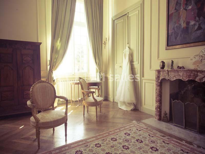 Robe de mariée suspendue dans une pièce élégante avec meubles anciens, cheminée et grandes fenêtres.