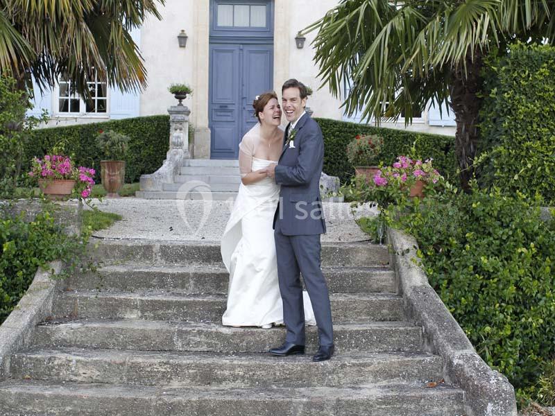 Un couple en tenue de mariage sourit sur les marches d'un escalier en pierre devant une maison entourée de végétation.