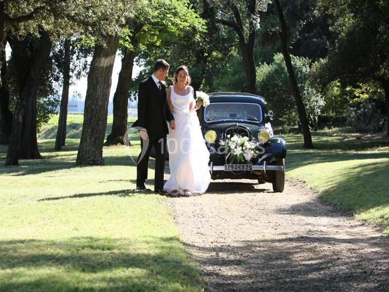 Un couple en tenue de mariage marche sur un chemin de terre, avec une voiture ancienne décorée en arrière-plan.