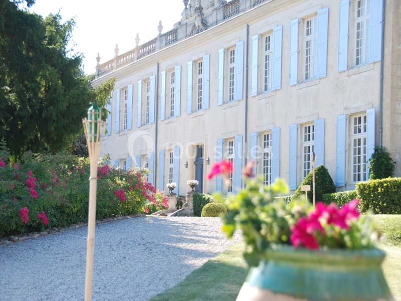 Façade d'un grand bâtiment ancien avec volets bleus, jardin fleuri et allée gravillonnée au premier plan.