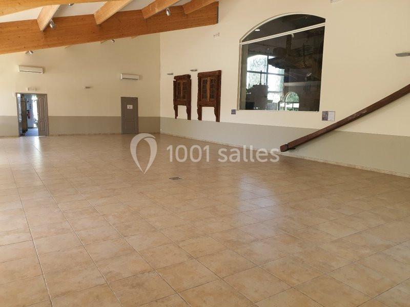 Salle spacieuse avec sol en carrelage beige, murs clairs, poutres apparentes et grandes fenêtres laissant entrer la lumière.