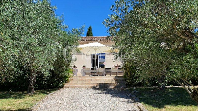 Maison avec terrasse ombragée par un parasol, entourée d'oliviers et d'un chemin en gravier sous un ciel bleu.