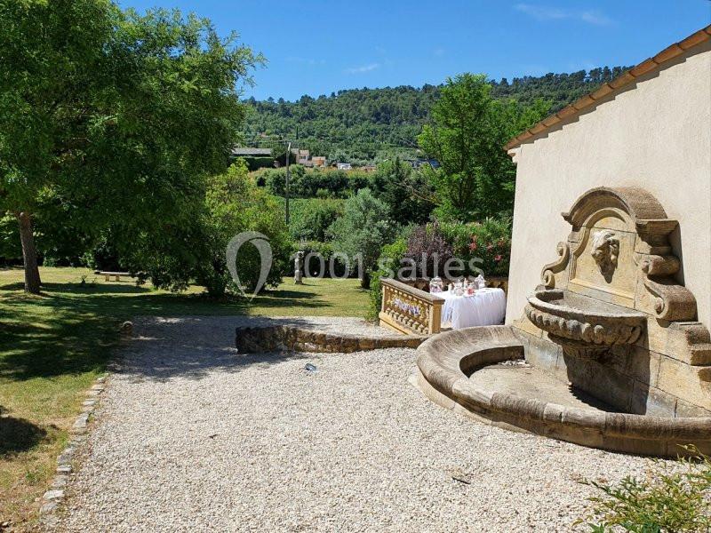 Cour en gravier avec une fontaine en pierre, entourée de verdure et d'arbres, sous un ciel bleu clair.
