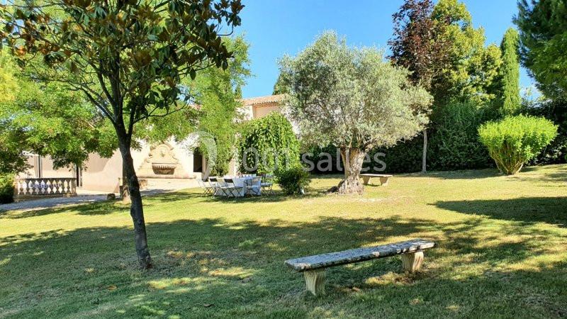 Jardin arboré avec banc en pierre, table et chaises blanches, fontaine et maison en arrière-plan.