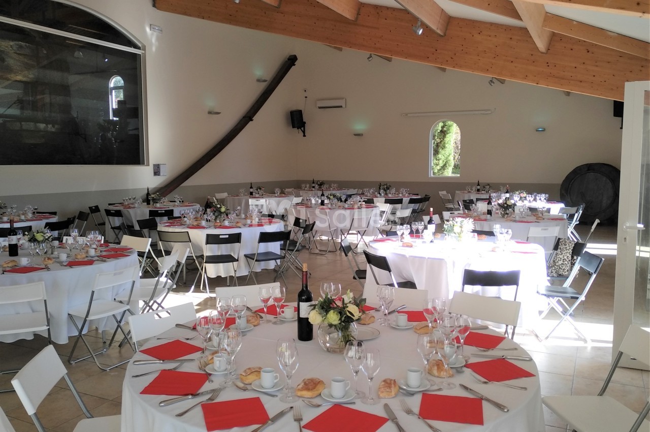 Salle de réception lumineuse avec tables rondes dressées, nappes blanches, serviettes rouges et décoration florale.