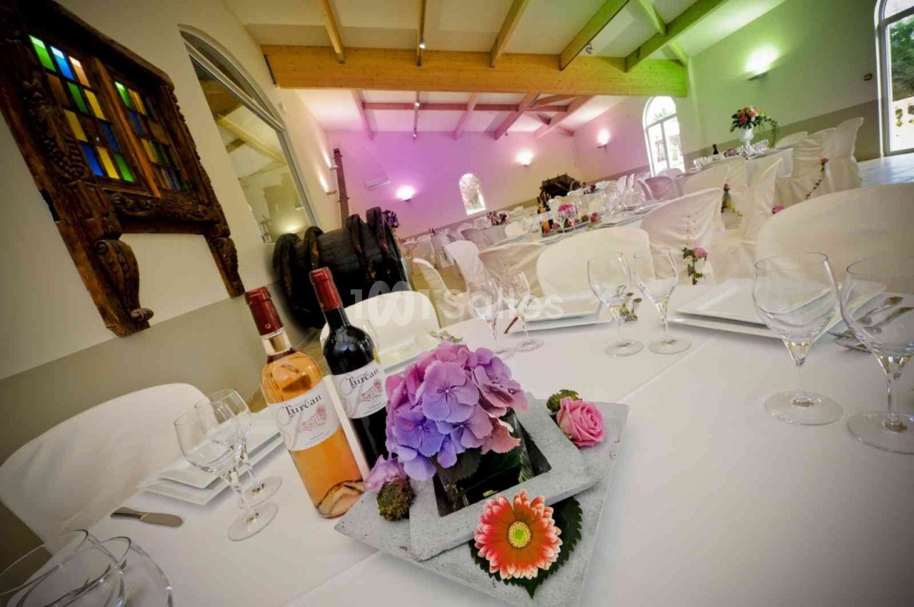 Salle de réception décorée avec des tables dressées, des fleurs en centre de table et des bouteilles de vin.
