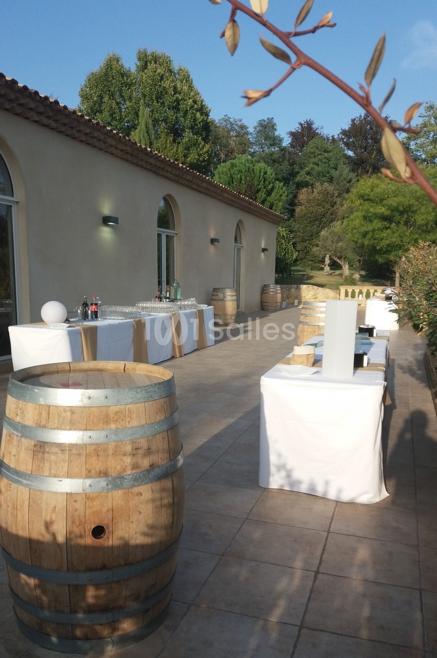 Terrasse ensoleillée avec tables dressées, tonneaux en bois et vue sur un jardin arboré.