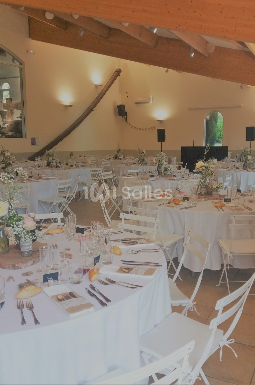 Salle de réception lumineuse avec tables rondes dressées, nappes blanches, chaises pliantes et décorations florales.