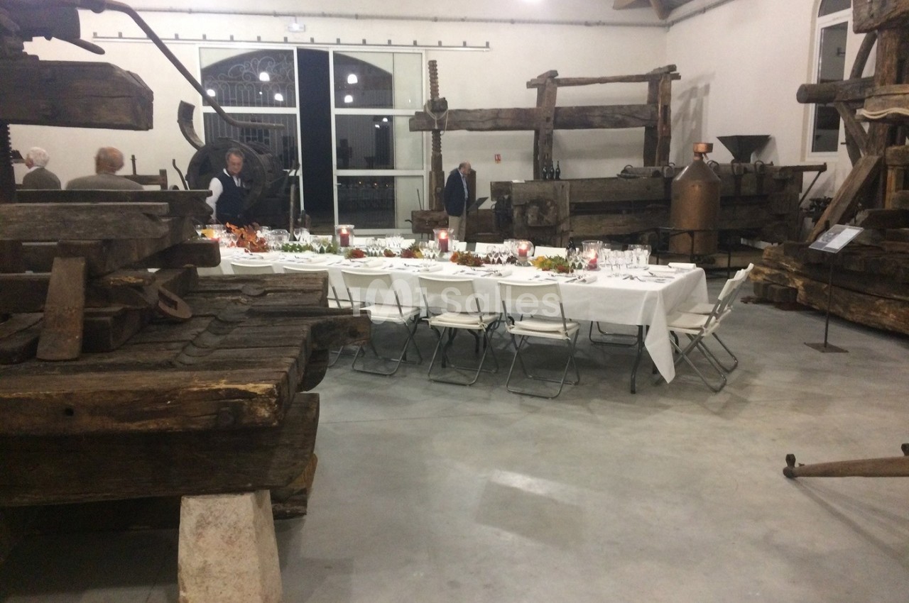 Salle avec des pressoirs anciens, une longue table dressée pour un repas, et quelques personnes en arrière-plan.