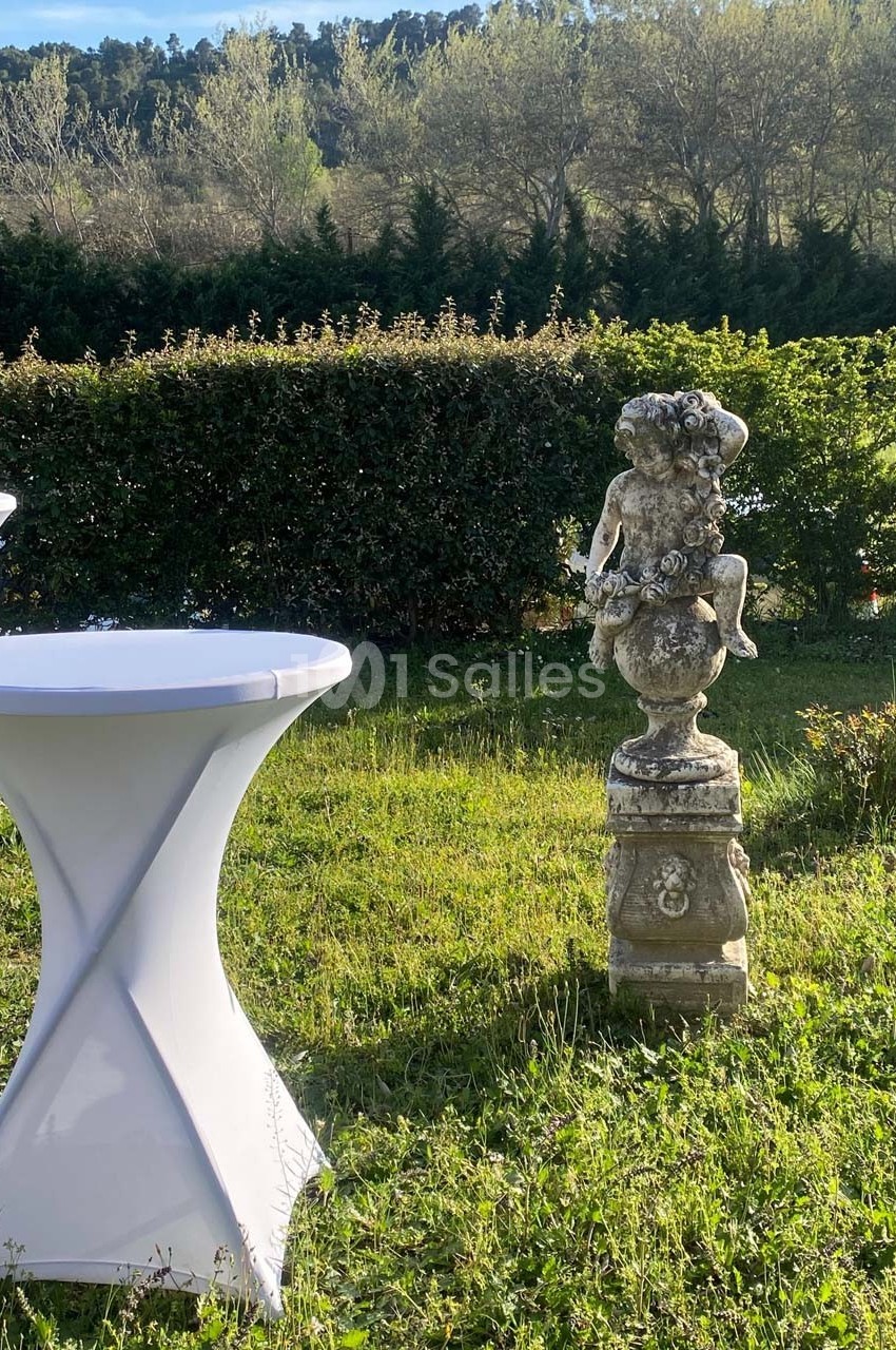 Statue en pierre représentant un enfant assis sur un piédestal, entourée de tables hautes et de verdure.