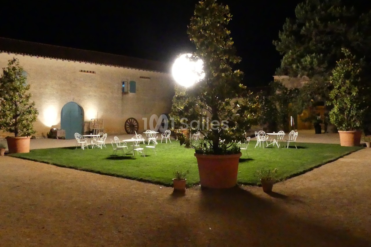 Cour extérieure éclairée la nuit, avec des tables et chaises blanches disposées autour d'une pelouse centrale.