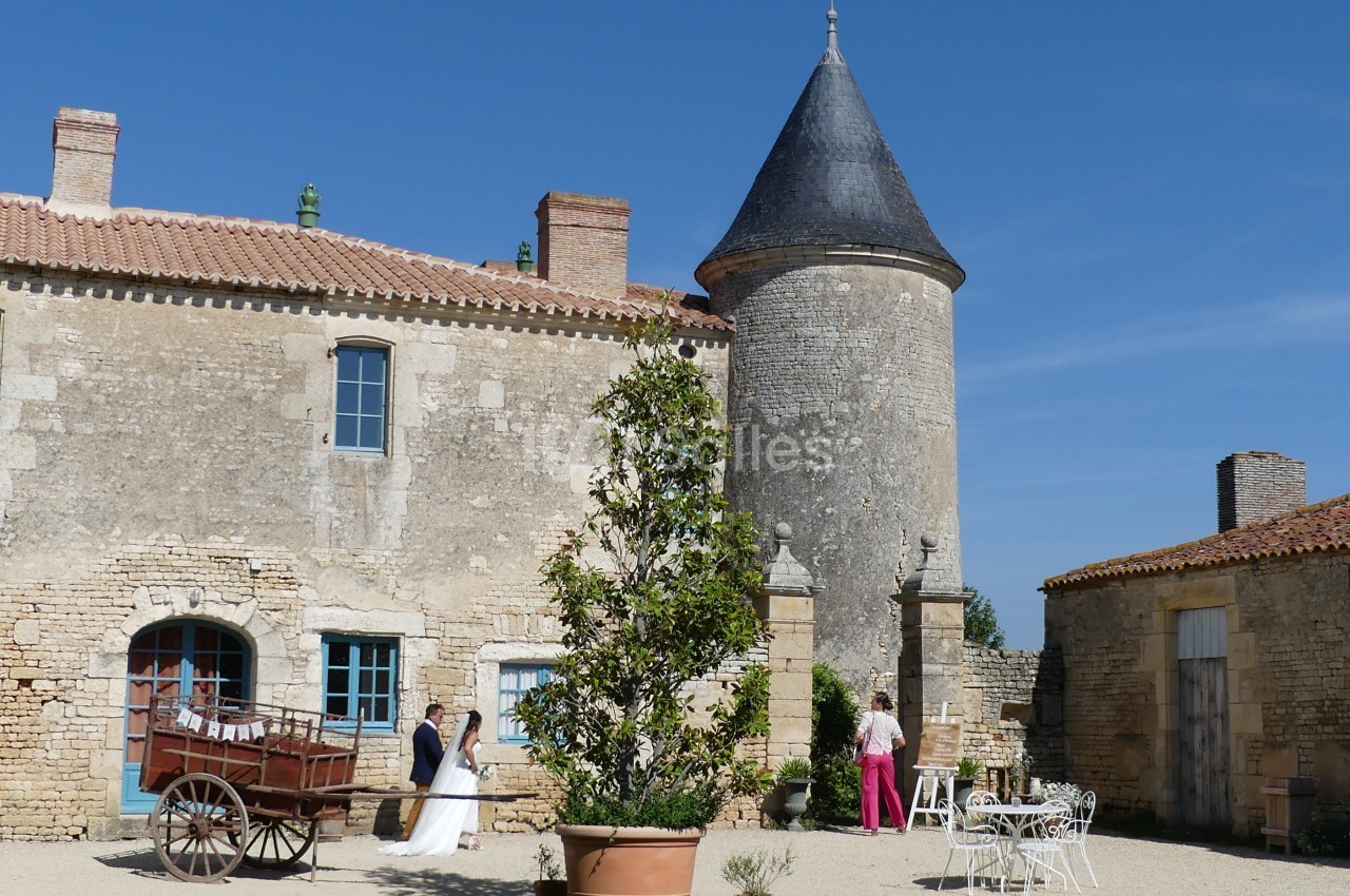 Mariés discutant dans la cour d'un château en pierre avec une tour ronde et des meubles de jardin blancs.