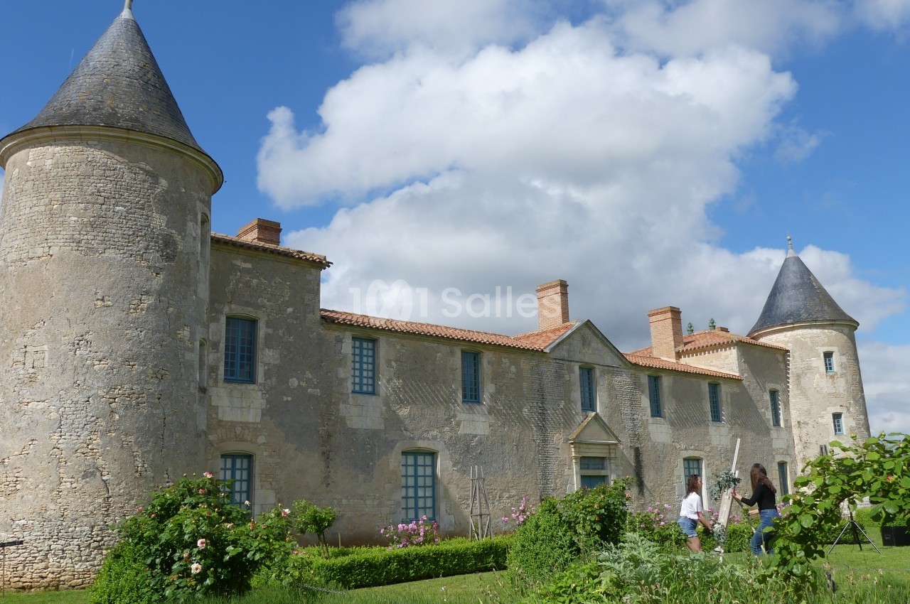 Façade d'un château en pierre avec tours rondes, fenêtres à volets bleus et jardin verdoyant au premier plan.