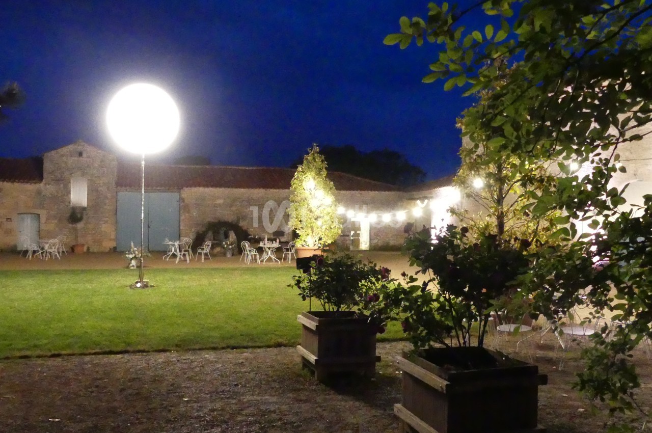 Cour extérieure éclairée de nuit avec des guirlandes lumineuses, des tables et des plantes en pot.