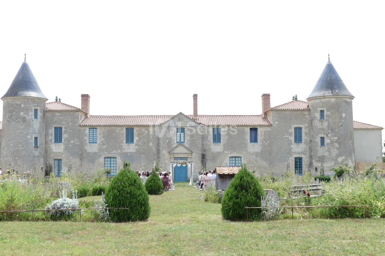 Façade d'un château ancien avec deux tours, entouré de jardins verdoyants et d'arbustes.