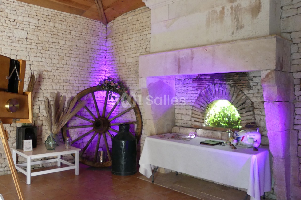 Salle en pierre avec une cheminée, une roue en bois décorative, une table blanche et un éclairage violet.