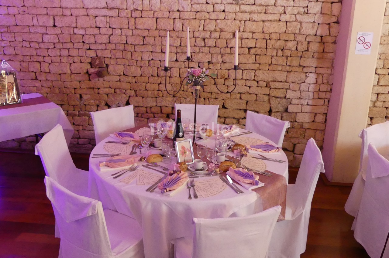 Table ronde décorée pour un événement, avec nappes blanches, chandeliers, menus et couverts, devant un mur en pierre.