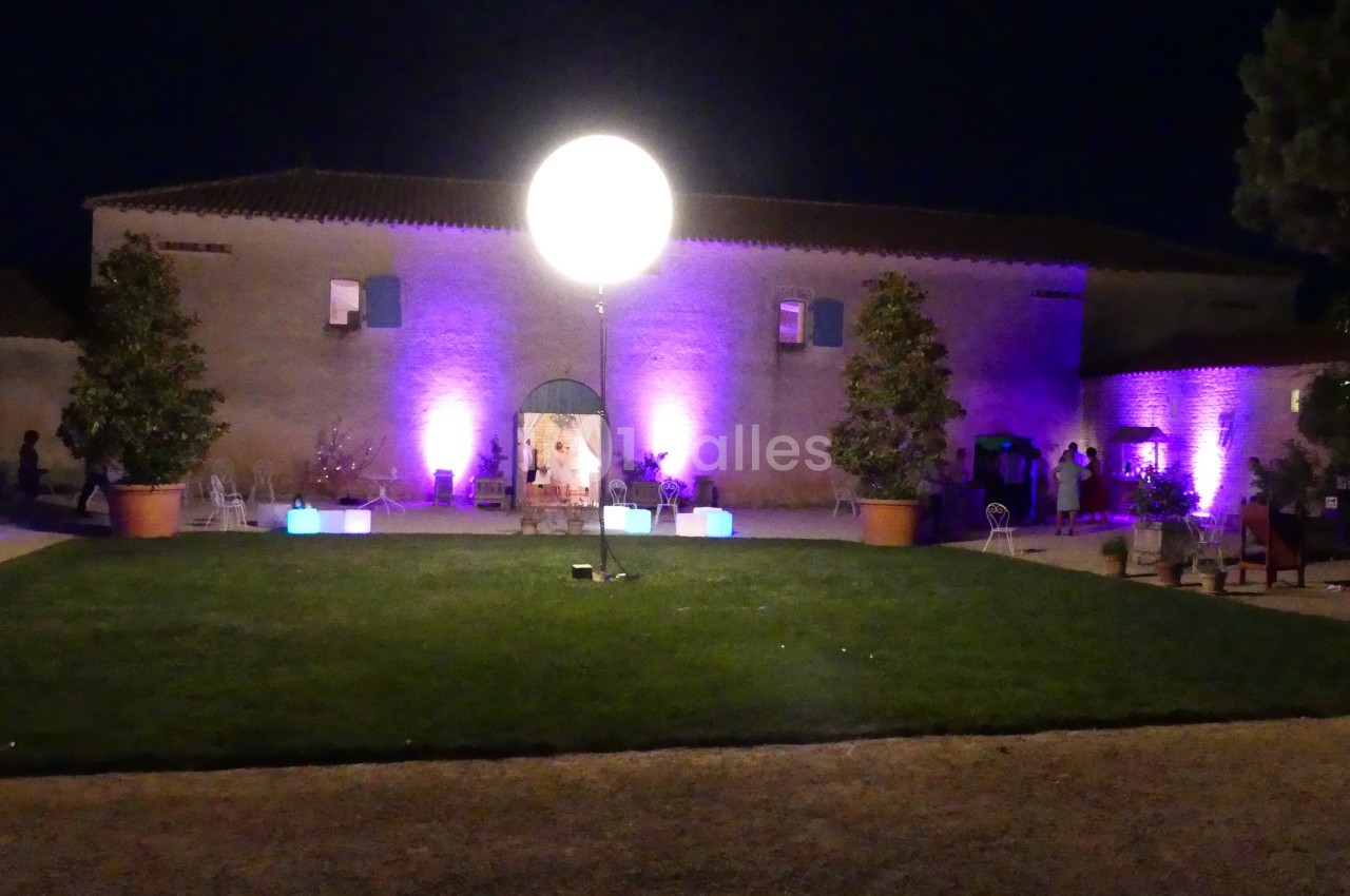 Cour extérieure éclairée la nuit avec un grand ballon lumineux central, des murs en pierre et des lumières colorées.