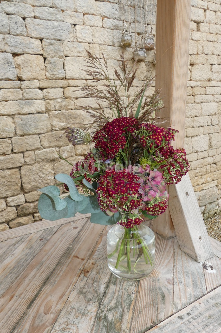 Bouquet de fleurs rouges et roses avec des feuillages dans un vase en verre, posé sur une table en bois devant un mur en…