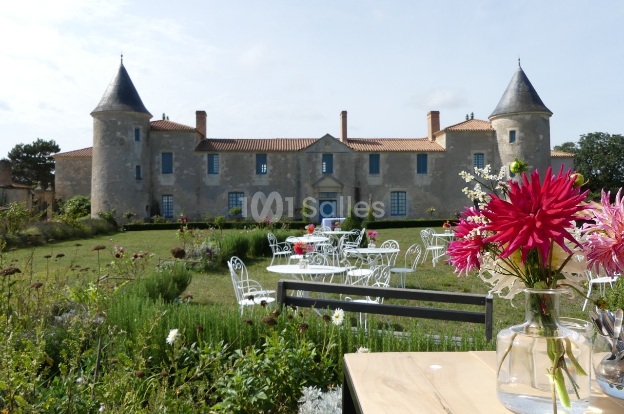 Façade d'un château avec tours, jardin fleuri, mobilier de jardin blanc et vase de fleurs sur une table au premier plan.