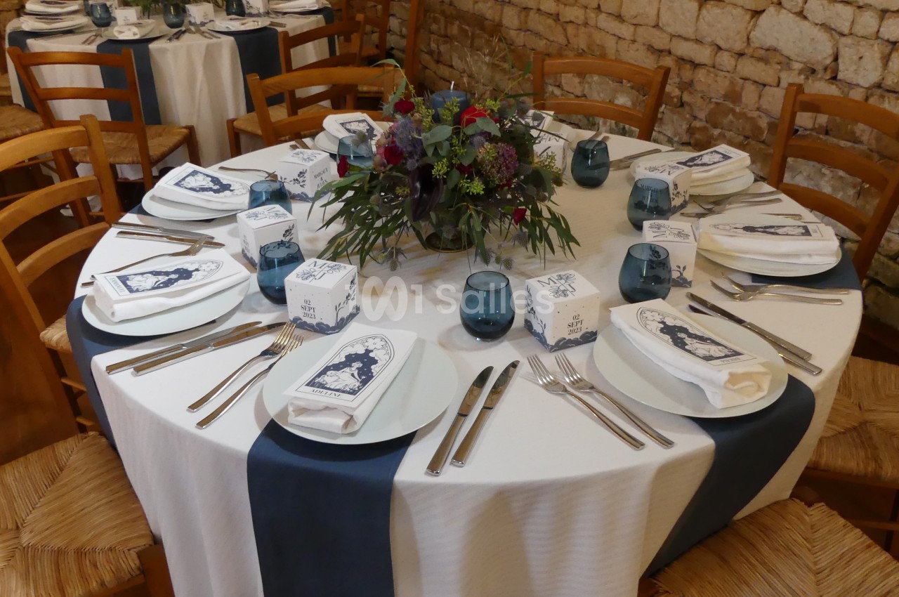 Table dressée avec nappes blanches et chemins de table bleus, ornée d'un centre de table floral et de couverts disposés.