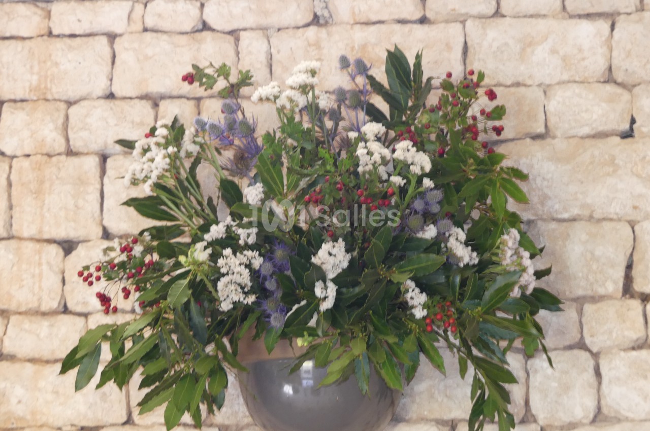 Arrangement floral coloré avec des fleurs blanches, rouges et violettes, suspendu devant un mur en pierre beige.