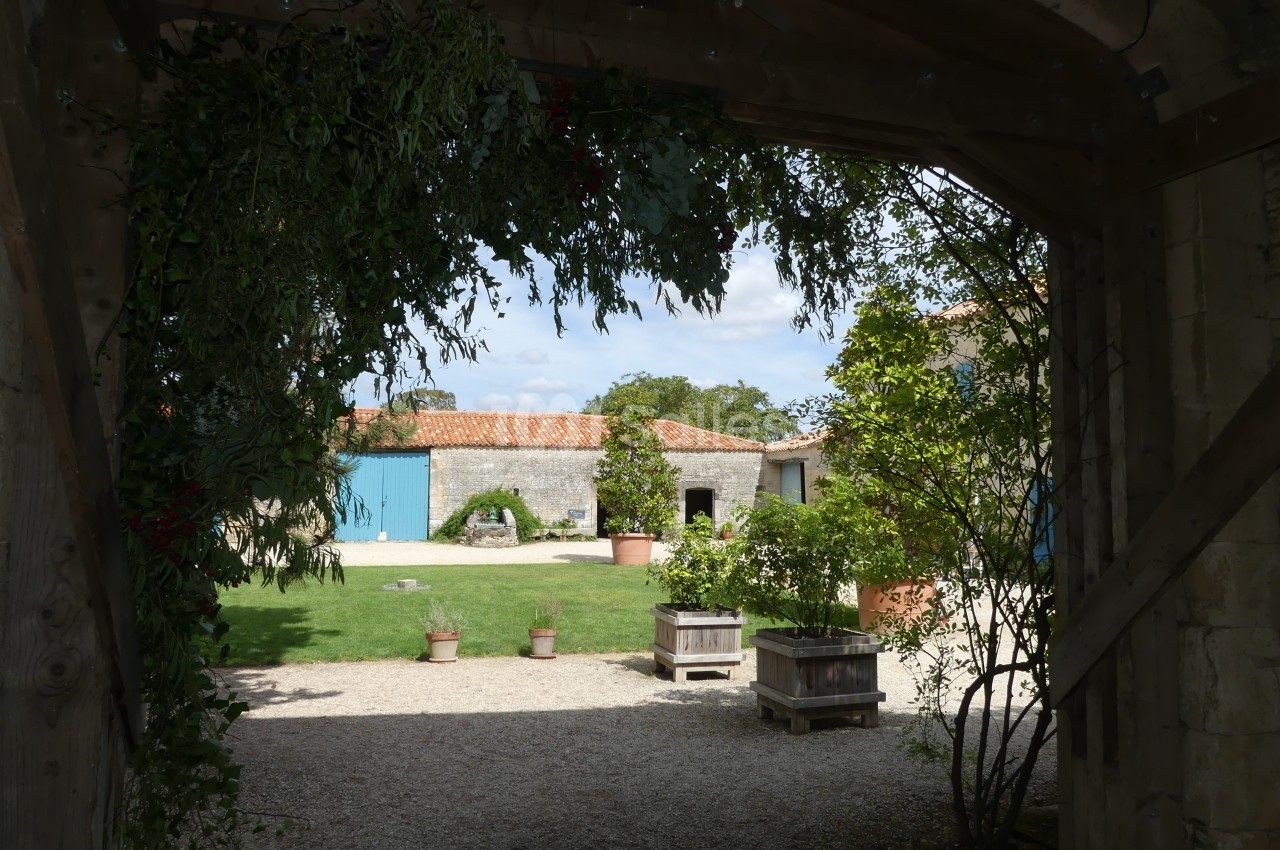 Vue d'une cour intérieure avec bâtiments en pierre, toits en tuiles, pelouse et jardinières sous une arche en bois.