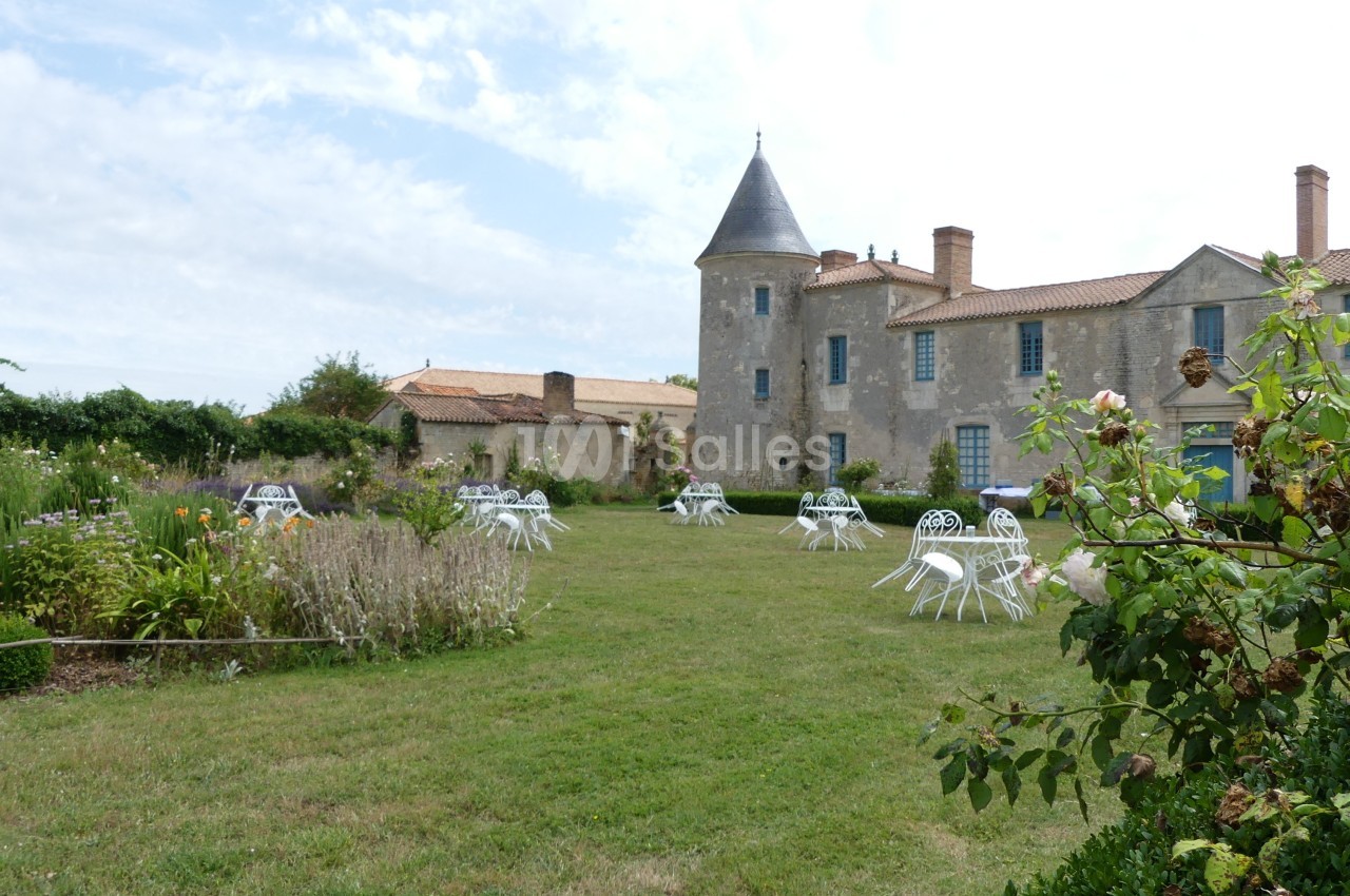 Jardin avec tables et chaises blanches devant un bâtiment ancien en pierre avec une tour et un toit en tuiles.