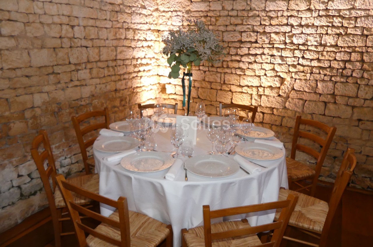 Table ronde dressée avec nappes blanches, vaisselle et bouquet central, entourée de chaises en bois, devant un mur en pierre.