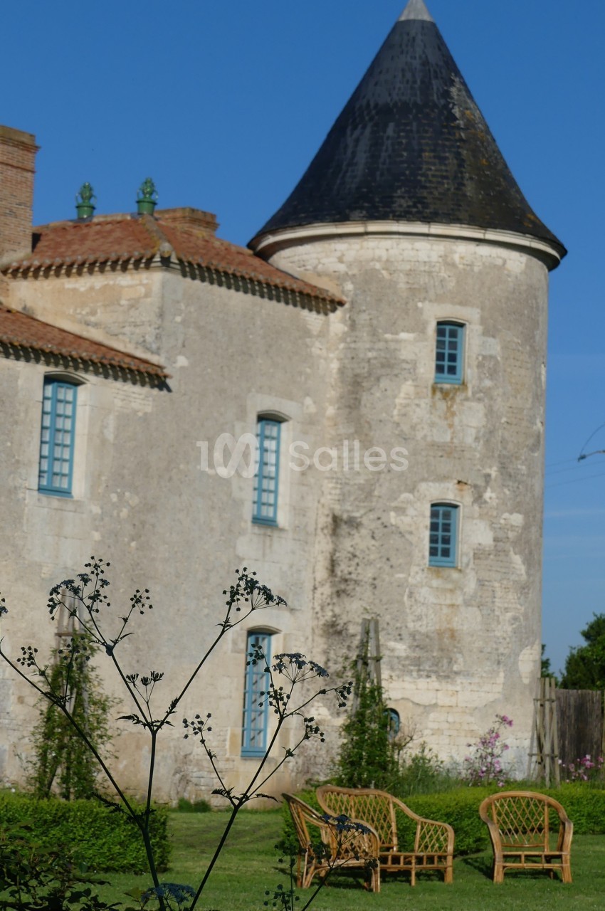 Tour en pierre d'un bâtiment ancien avec des fenêtres bleues, entourée de verdure et de mobilier de jardin en rotin.