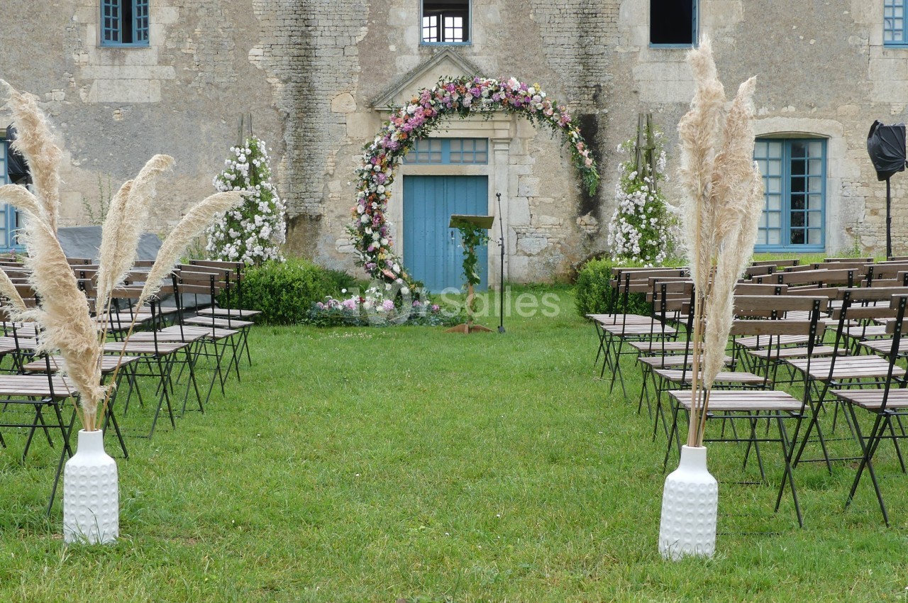 Allée centrale d'une cérémonie en plein air, décorée d'herbes de la pampa et d'une arche fleurie devant un bâtiment ancien.